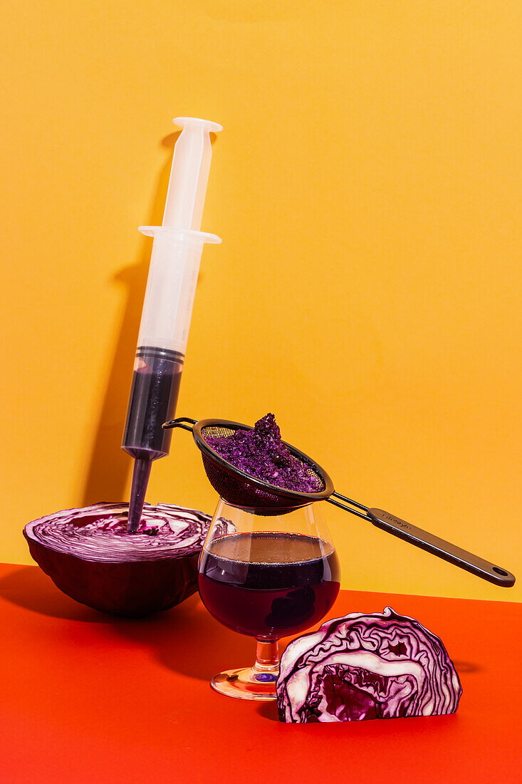 Composition of glass with strainer and syringe filled with homemade juice of red cabbage pricked in cabbage against bright background