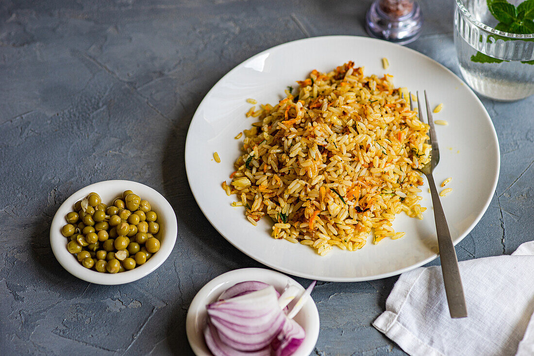 High angle of boiled rice with onion, carrot, bell pepper, green peas and spices on plate with fork near slices of onion and glass of water with mint against gray background