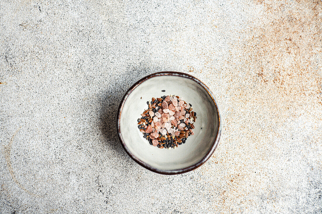 Schüssel mit Salatgewürzmischung bestehend aus rosa Himalaya-Salz, Leinsamen, Sesam und schwarzem Pfeffer vor einem Betonhintergrund
