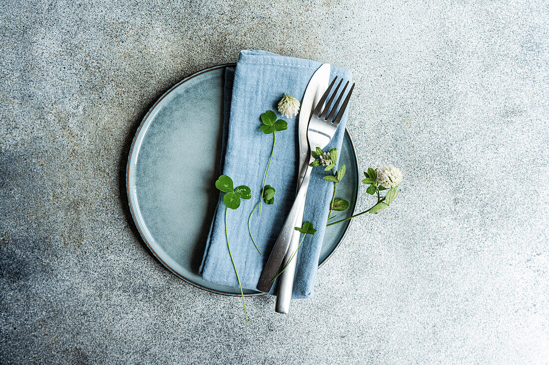 Draufsicht auf einen gedeckten Tisch mit Blättern einer Kleeblattpflanze auf einem Teller mit Serviette, Gabel und Messer vor einem Hintergrund aus Beton