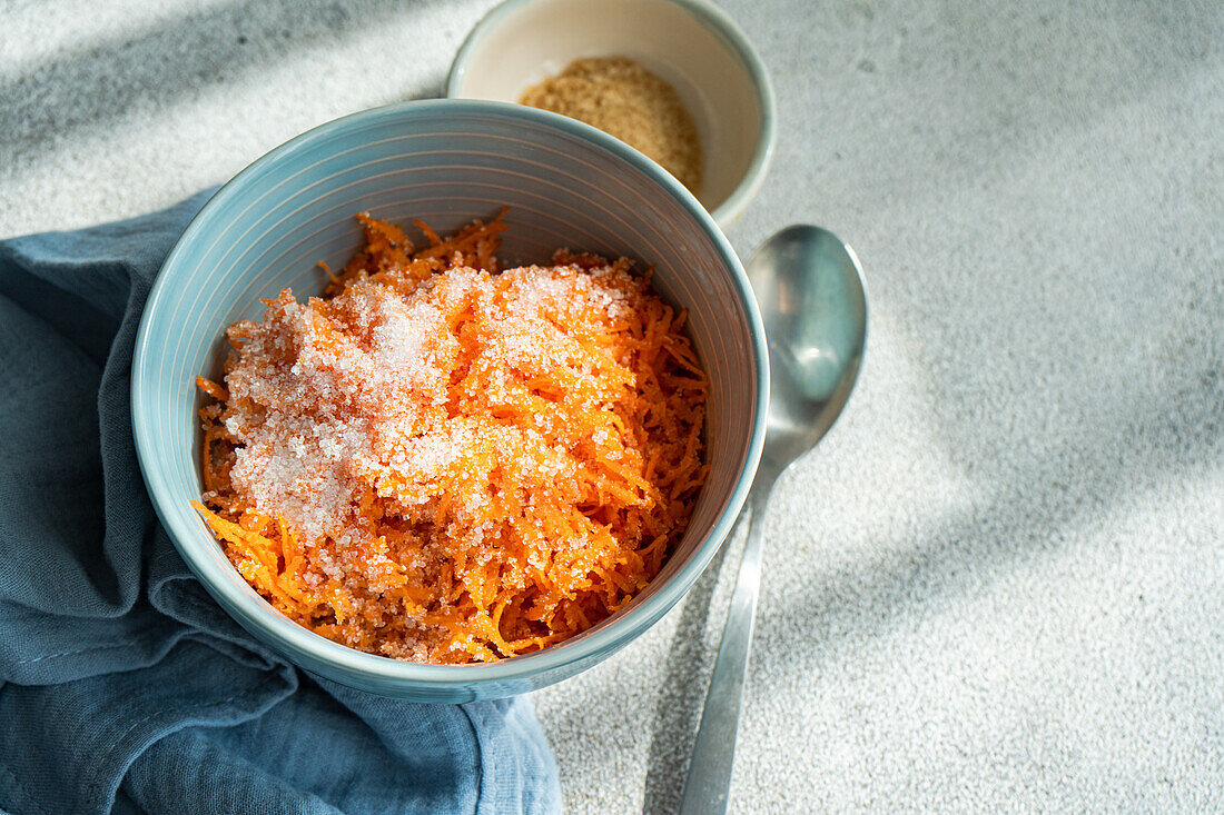 Top view of a light blue bowl containing freshly grated carrot sprinkled with sugar, placed next to a gray cloth, a small beige bowl with brown sugar, and a metallic spoon, all set against a textured gray background with sunlight casting soft shadows