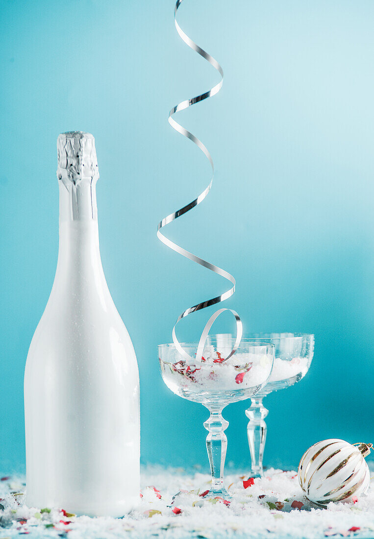 Christmas and New Year setting with champagne bottle, glasses, falling silver ribbon, snow and bauble and at light blue background. Front view