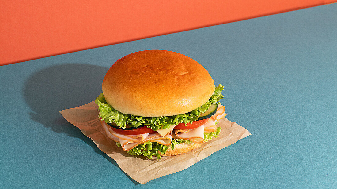 Leckeres Sandwich mit Schinken, Tomaten, Gurken- und Käsescheiben und frischem Salat auf blauem und orangem Hintergrund