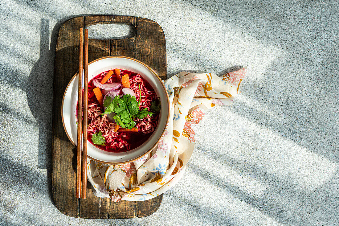 Schale mit Rote-Bete-Suppe mit Zwiebeln, Koriander und Nudeln im asiatischen Stil, serviert auf einer Schale und Stäbchen auf einer Serviette vor einer grauen Fläche im Tageslicht (Draufsicht)