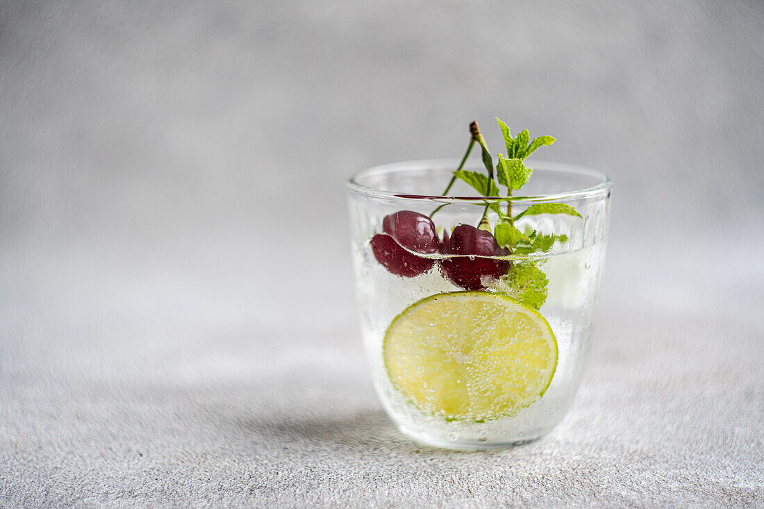 Transparent glass of refreshing cold drink with lime and mint leaves on gray surface in summer day against blurred background