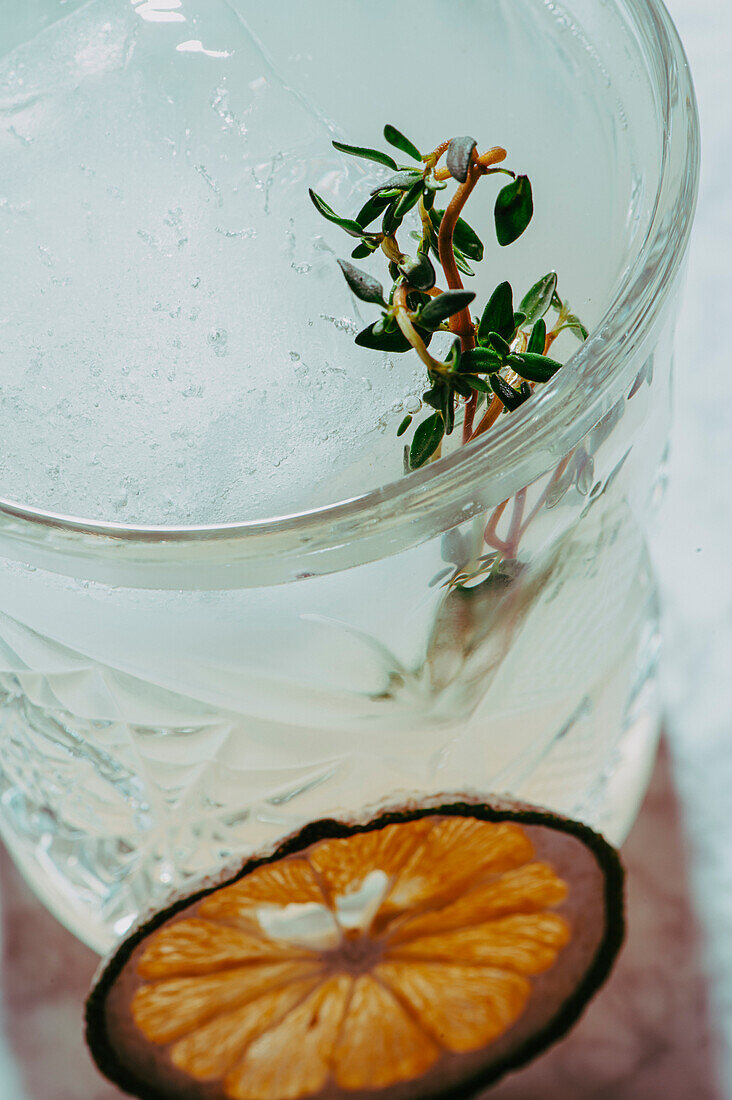Hoher Winkel eines aromatischen Cocktails mit Eiswürfeln und Kräutern in einem transparenten Glas, dekoriert mit einer getrockneten Zitrusscheibe