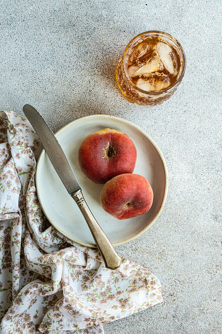 Draufsicht auf einen Teller mit einer Pfirsichsorte und einer Sommerlimonade mit Eis und Messer auf einer Serviette vor grauem Hintergrund