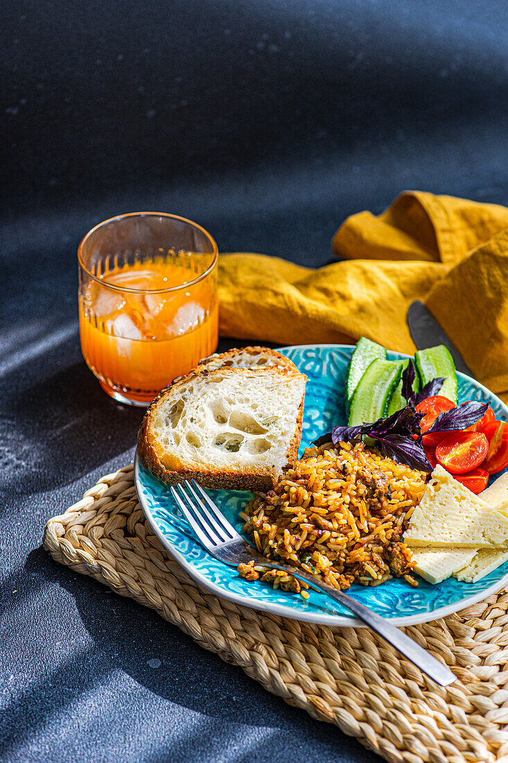 High angle of five ingredients lunch set consists of homemade goat cheese, fresh cucumber and tomato with leaves of basil herb, in addition to fried rice with meat with slices of healthy sour dough bread served with glass of carrot-apple juice against gray background