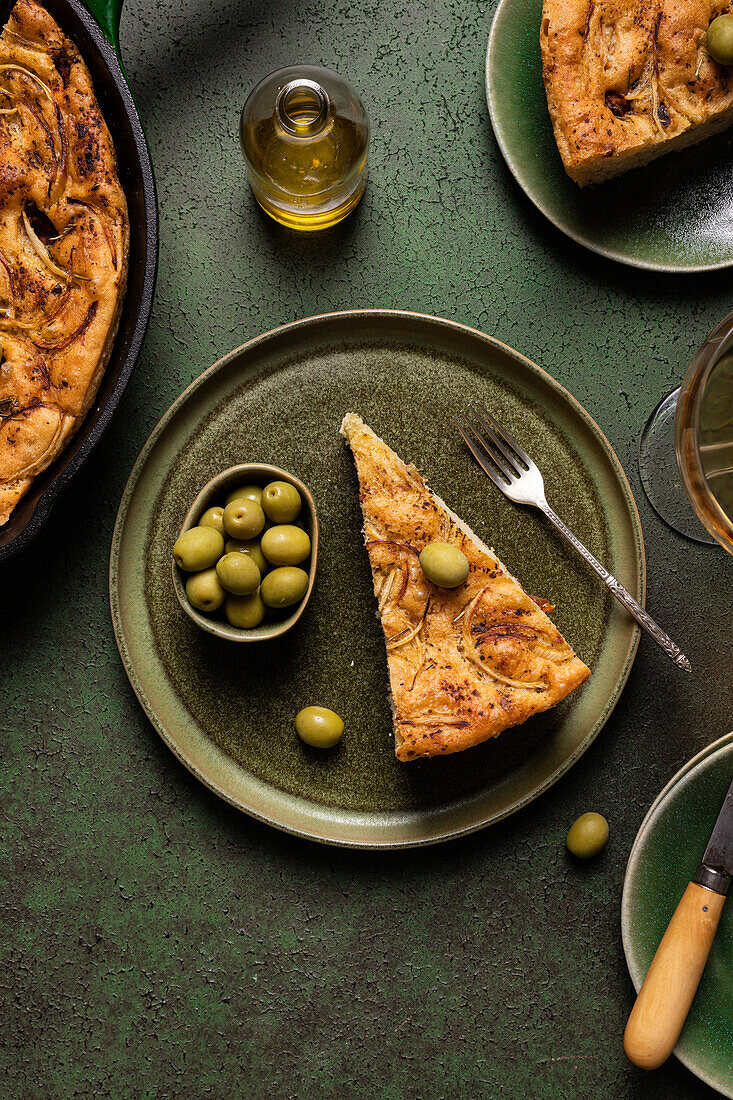 Top view of slice baked Italian focaccia bread with green olives in plate on dining table with drink on green background