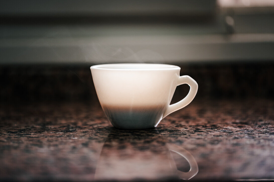 Eine dampfende weiße Kaffeetasse steht elegant auf einer Granitarbeitsplatte, die von oben mit sanftem Umgebungslicht beleuchtet wird und ein Gefühl von Ruhe und Morgenritual ausstrahlt