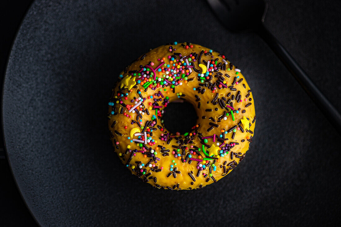 Top view of sweet banana donut with colorful sprinkles placed on black plate against dark surface