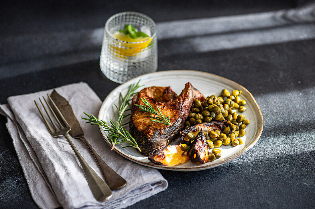 From above grilled salmon steak served with fermented capers and a wedge of lemon garnished with fresh rosemary on a rustic ceramic plate with vintage cutlery