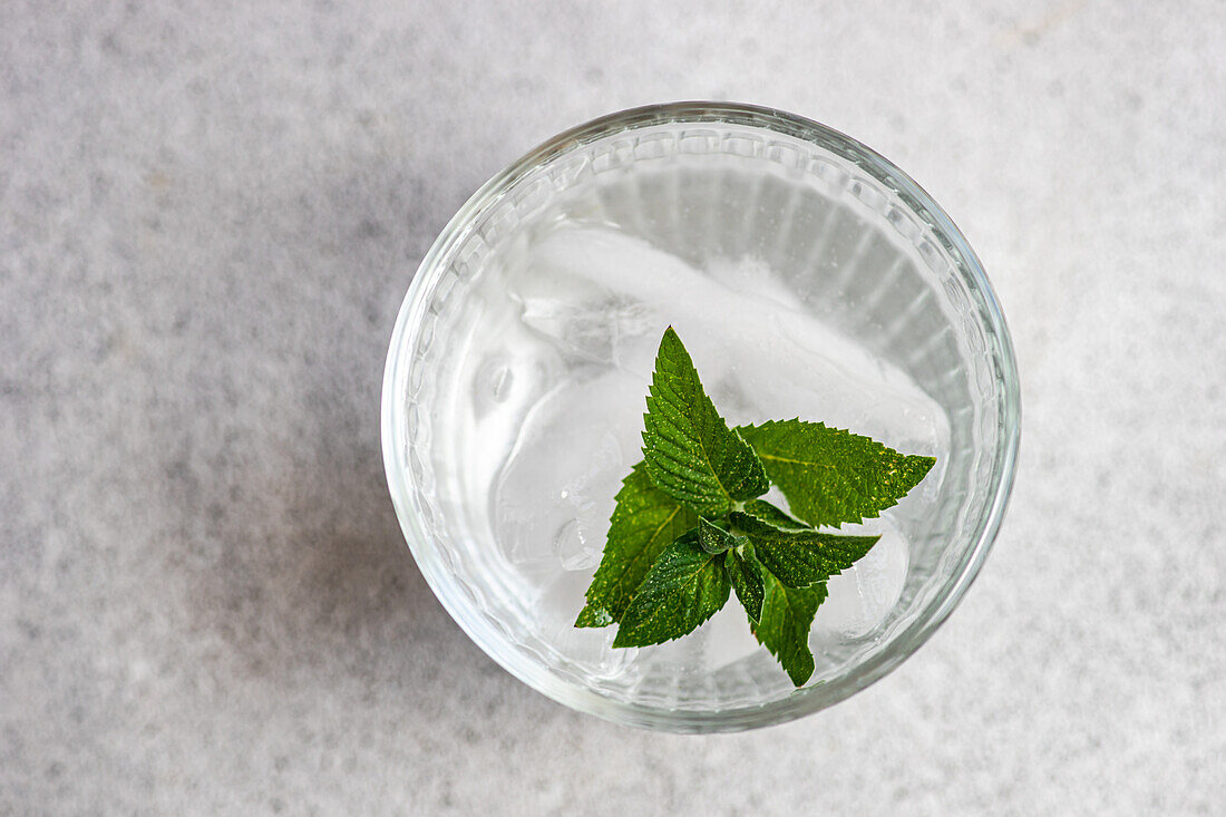Draufsicht auf ein Glas Mineralwasser mit Eiswürfeln und frischen Minzblättern vor grauem Hintergrund