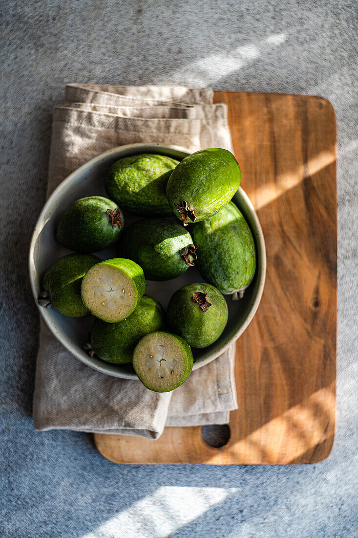 A bowl of ripe feijoa fruits, some cut in half, beautifully presented on a wooden kitchen board with a linen napkin under natural lighting.