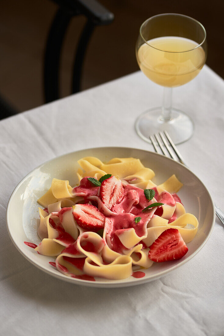 Ein köstliches Nudelgericht mit Erdbeerrahmsauce, garniert mit frischen Erdbeerscheiben und Minze auf einem stilvollen Teller neben einem Glas Weißwein
