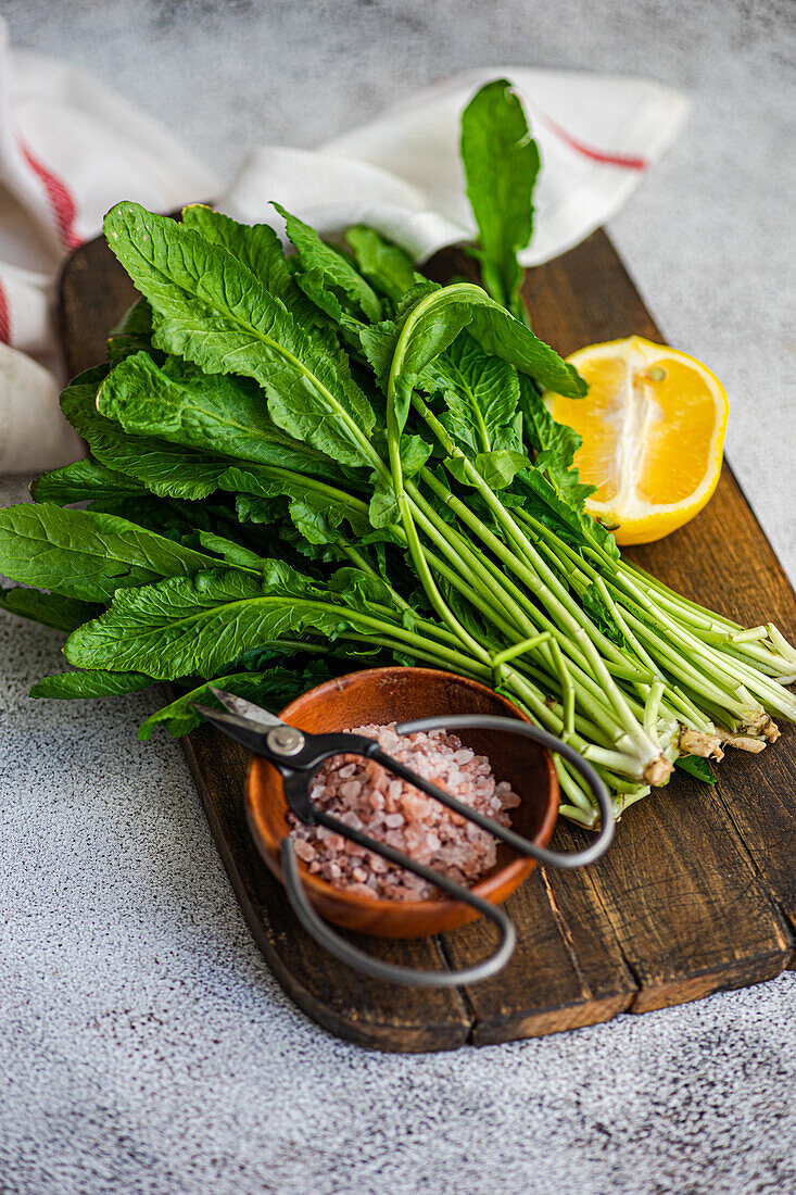 From above of fresh arugula leaves with a slice of lemon and a bowl of salt on a wooden cutting board, accented by kitchen scissors and a striped towel