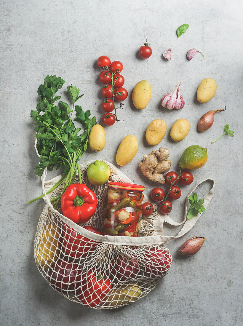 Sustainable groceries shopping bag with various vegetables: potatoes, tomatoes, pepper, lime, herbs, garlic, onion and preserve on grey concrete table. Plastic free lifestyle . Top view. Zero waste