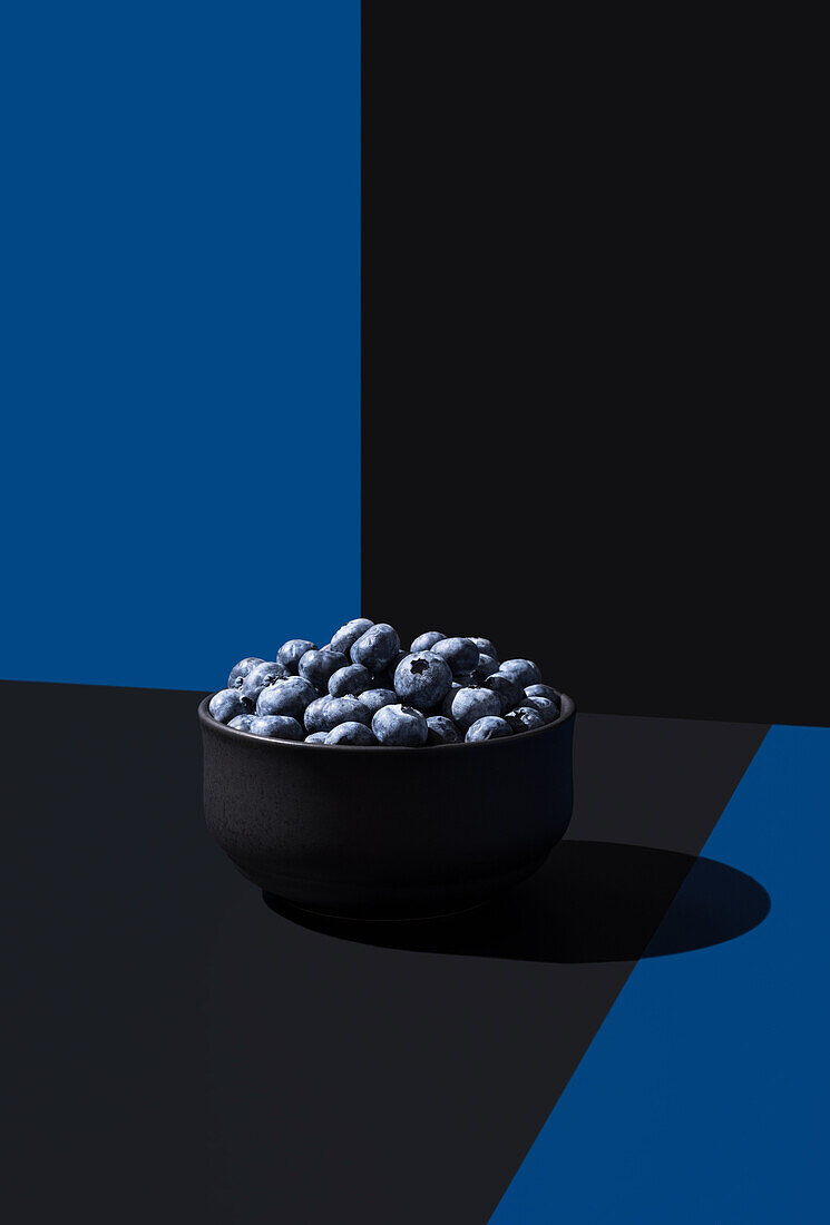Eine minimalistische Komposition mit einer schwarzen Schale Blaubeeren auf einem geteilten blauen und schwarzen Hintergrund mit dramatischer Beleuchtung