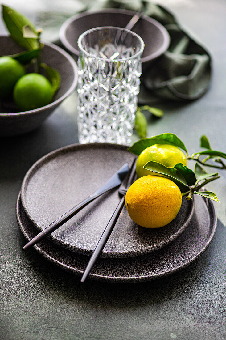 A sophisticated table setting featuring black cutlery on a dark plate with a bright yellow lemon, alongside a textured glass and fresh limes.