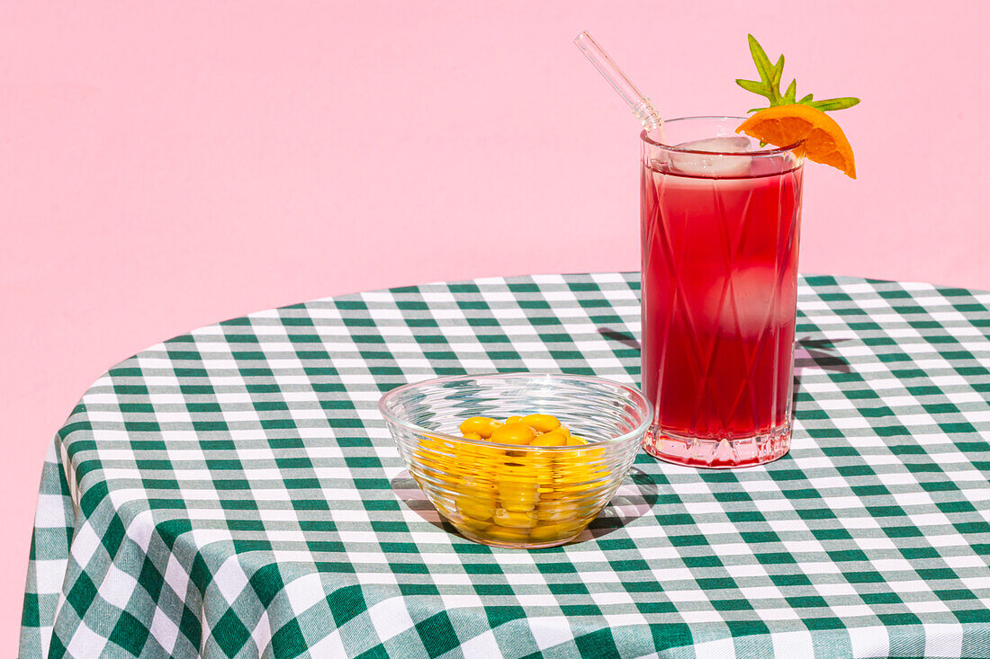 Glass of cold refreshing red cocktail with ice and straw served with orange near transparent bowl with lupin bean on table with checkered tablecloth