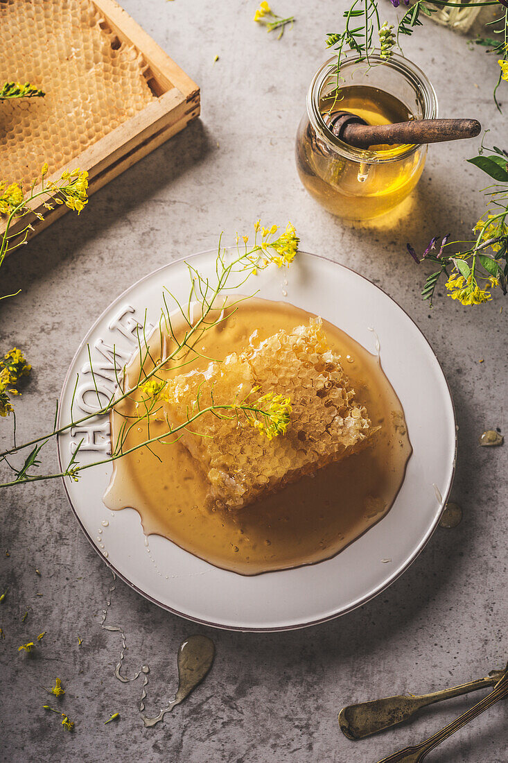 Whole raw honeycomb on table with honey jar and wooden dipper, yellow flowers with petals. Healthy and natural liquid sweetener on grey concrete kitchen table. Top view.