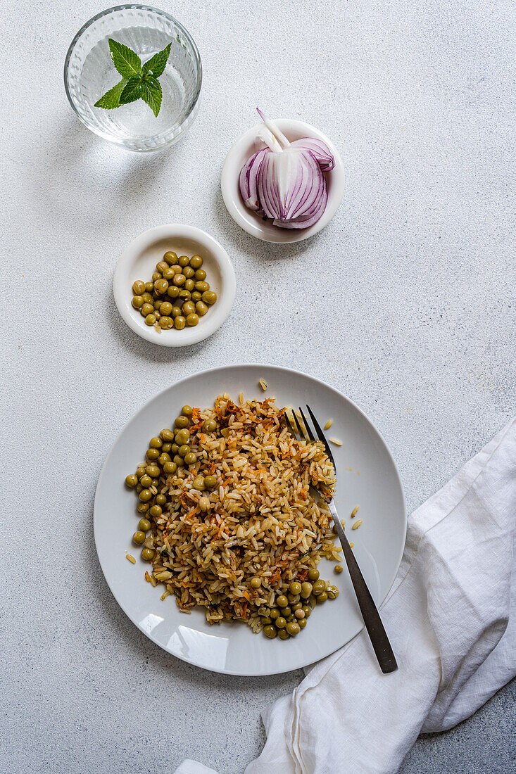 Draufsicht auf gekochten Reis mit Zwiebel, Karotte, Paprika, grünen Erbsen und Gewürzen auf einem Teller mit Gabel neben Zwiebelscheiben und einem Glas Wasser mit Minze vor weißem Hintergrund