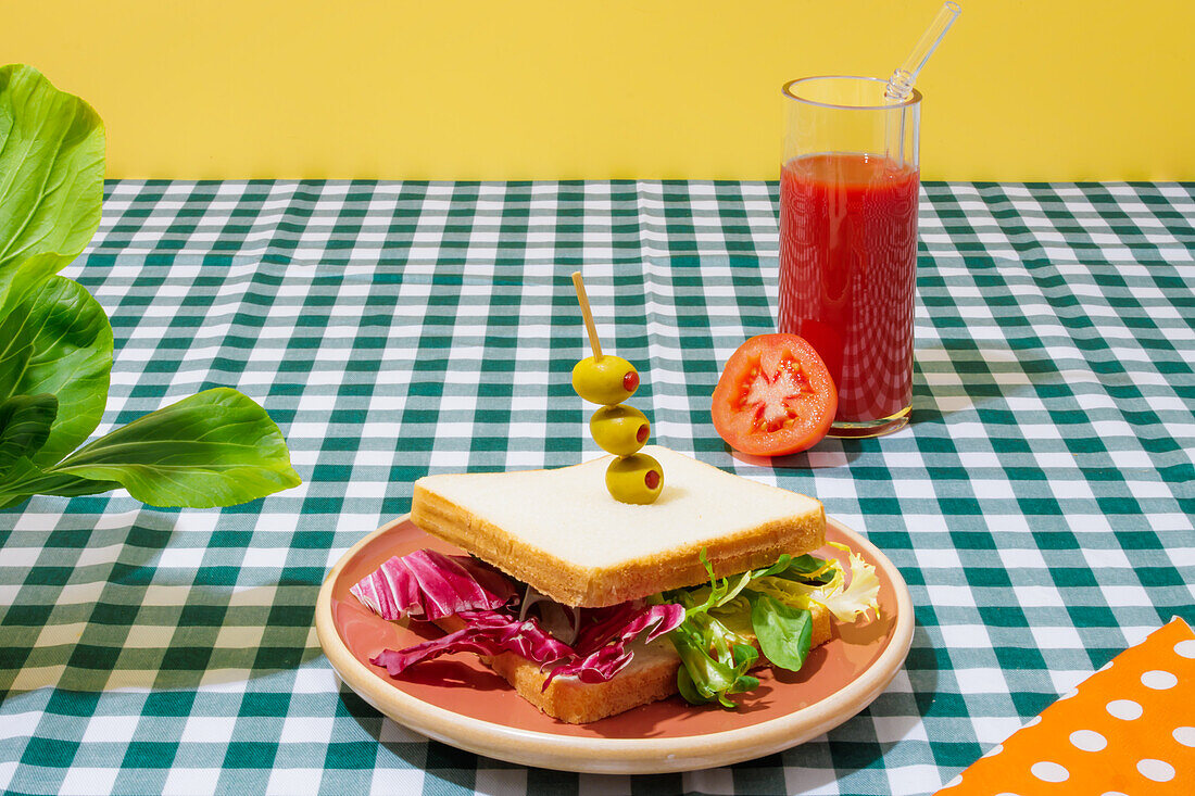 Appetitliches gesundes Sandwich mit frischem Salat auf einem Teller mit Oliven neben einem Glas Tomatensaft mit Glasstrohhalm auf einem karierten Tischtuch