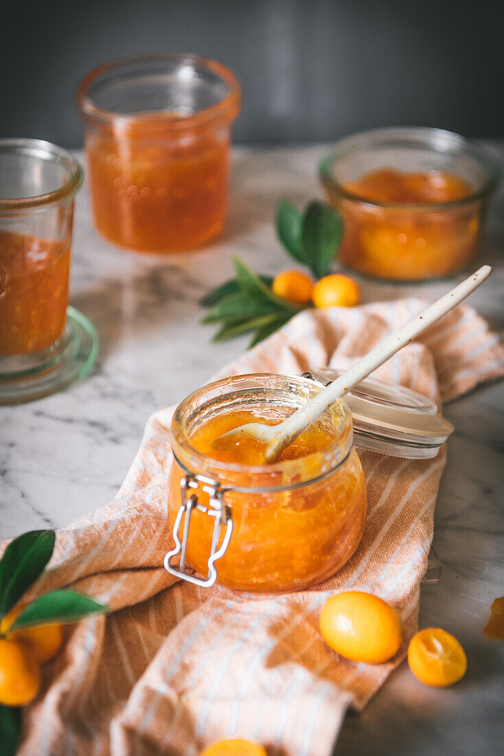 Nahaufnahme eines Glases mit frischer Kumquat-Marmelade mit Löffel auf einem Tisch vor verschwommenem dunkelgrauem Hintergrund