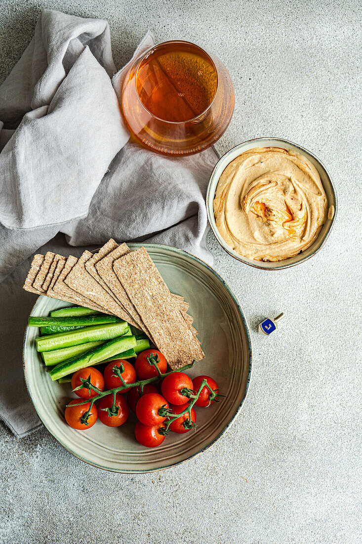 Top view of healthy plant-based plate with hummus and vegetables served in bowls near napkin and glass of liquor against gray background