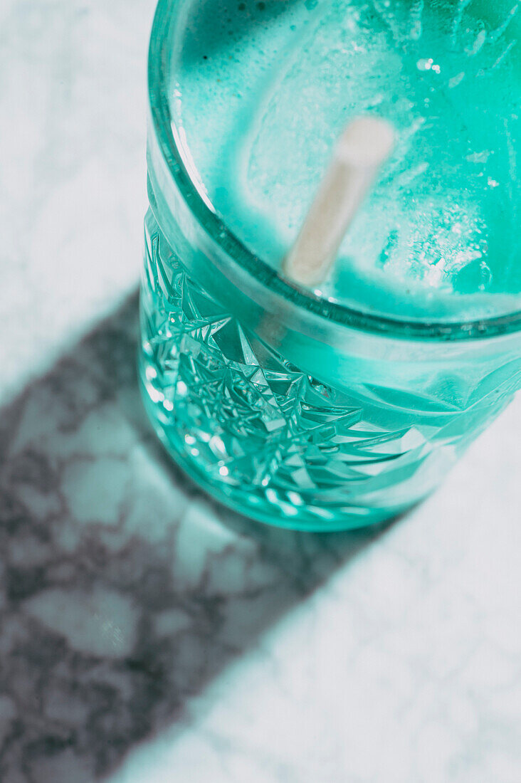 Hoher Winkel eines transparenten Glases, gefüllt mit einem bunten, erfrischenden, kalten Alkoholcocktail und Eiswürfeln mit Strohhalm auf einer Marmorfläche