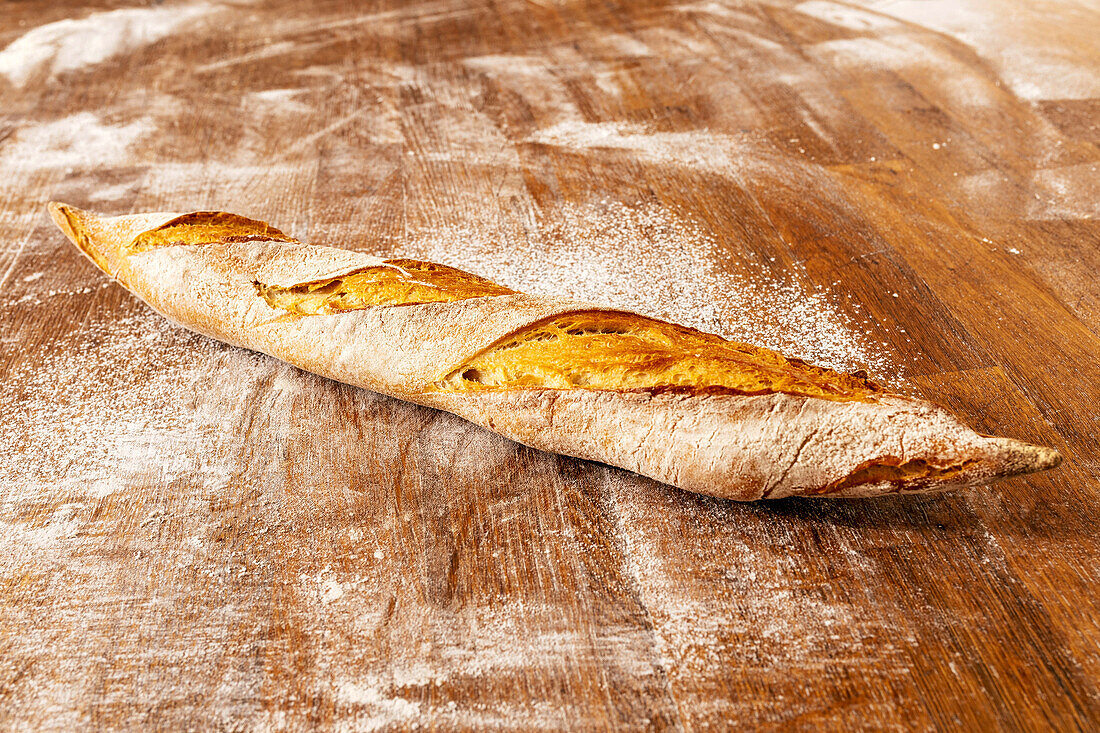 Closeup of freshly baked delicious baguette with crunchy crust placed on messy wooden table in kitchen at bakehouse