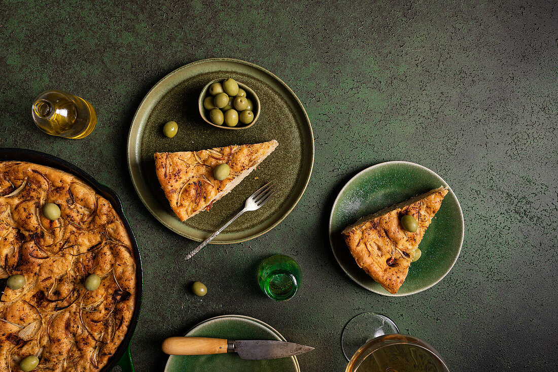 Draufsicht auf eine Scheibe gebackenes italienisches Focaccia-Brot mit grünen Oliven in einem Teller auf einem Esstisch mit Getränk auf grünem Hintergrund