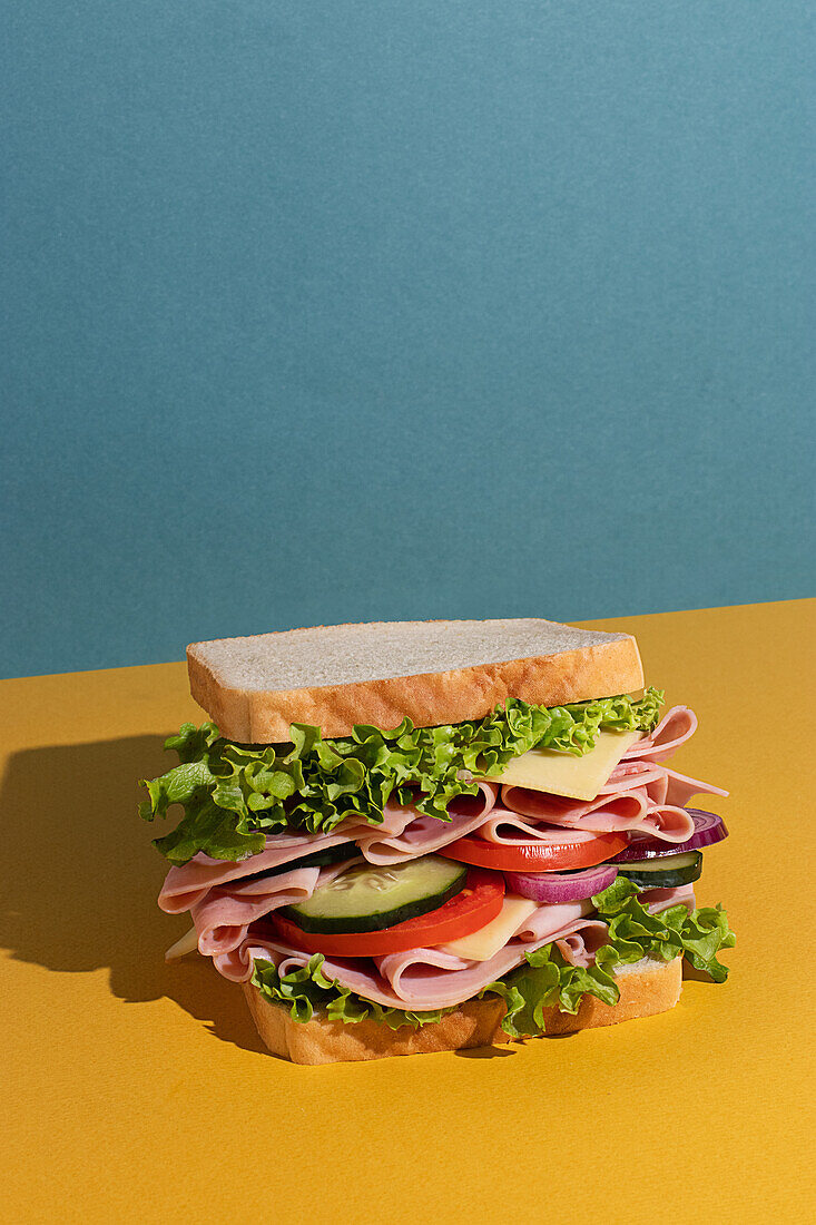 Leckeres Sandwich mit Schinken, Tomaten, Gurken- und Käsescheiben und frischem Salat auf blau-gelbem Hintergrund