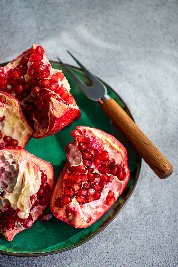 From above fresh pomegranate pieces with vibrant red seeds on a green ceramic plate set against a textured grey concrete background