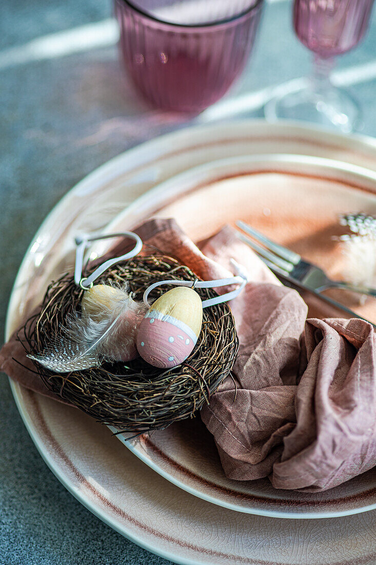 An artistic Easter table setting featuring a nest with pastel decorated eggs atop ceramic plates, complemented by a dusty pink napkin.