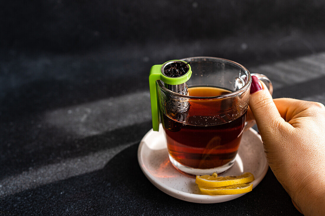 Anonyme Hand, die eine gläserne Teetasse mit grünem Teesieb und Zitronenscheiben auf einer Untertasse hält, vor einem dunklen Hintergrund