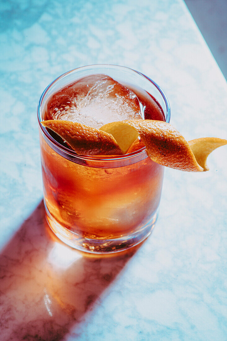 Blick von oben auf ein Glas mit einem altmodischen Cocktail und Eiswürfeln, die mit Orangenschalen verziert sind, auf einer Marmorfläche