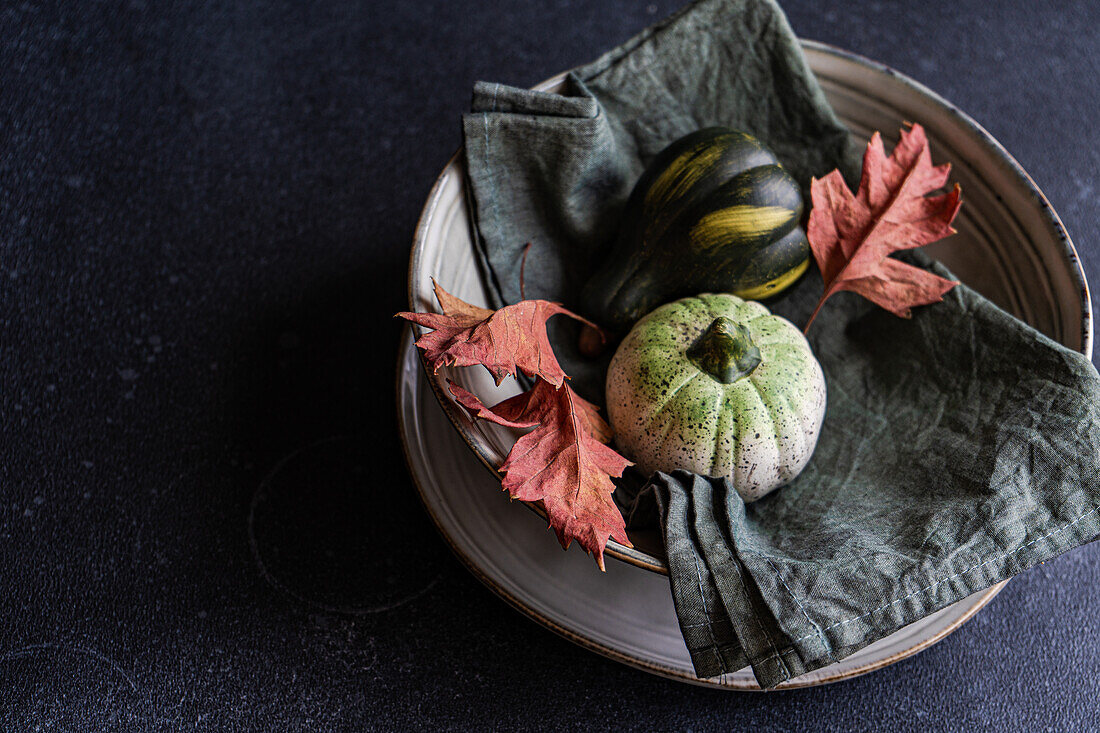High angle of autumnal table setting with napkin, leaves and pumpkins placed on ceramic plate against dark surface