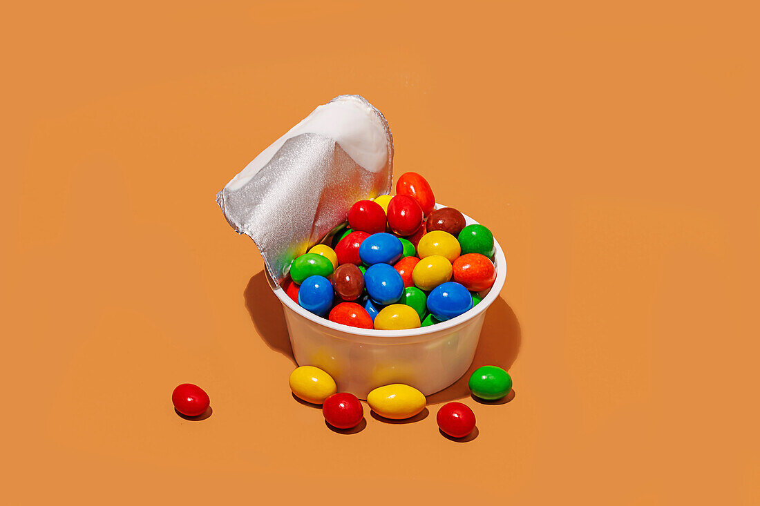 Hoher Winkel eines geöffneten Joghurtbehälters, gefüllt mit bunten Bonbons vor orangefarbenem Hintergrund