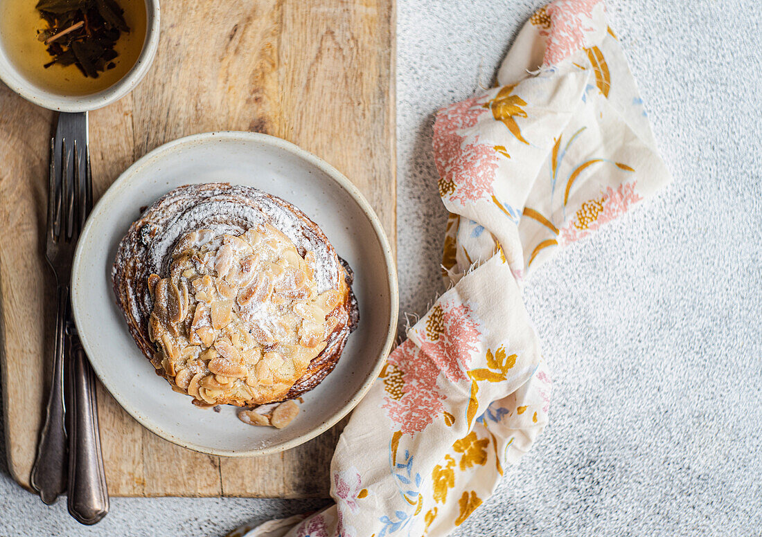 Draufsicht auf süßes Mandelgebäck auf Holzstäbchen zwischen Gabel und Messer und Serviette mit Tasse grünem Tee vor grauem Hintergrund