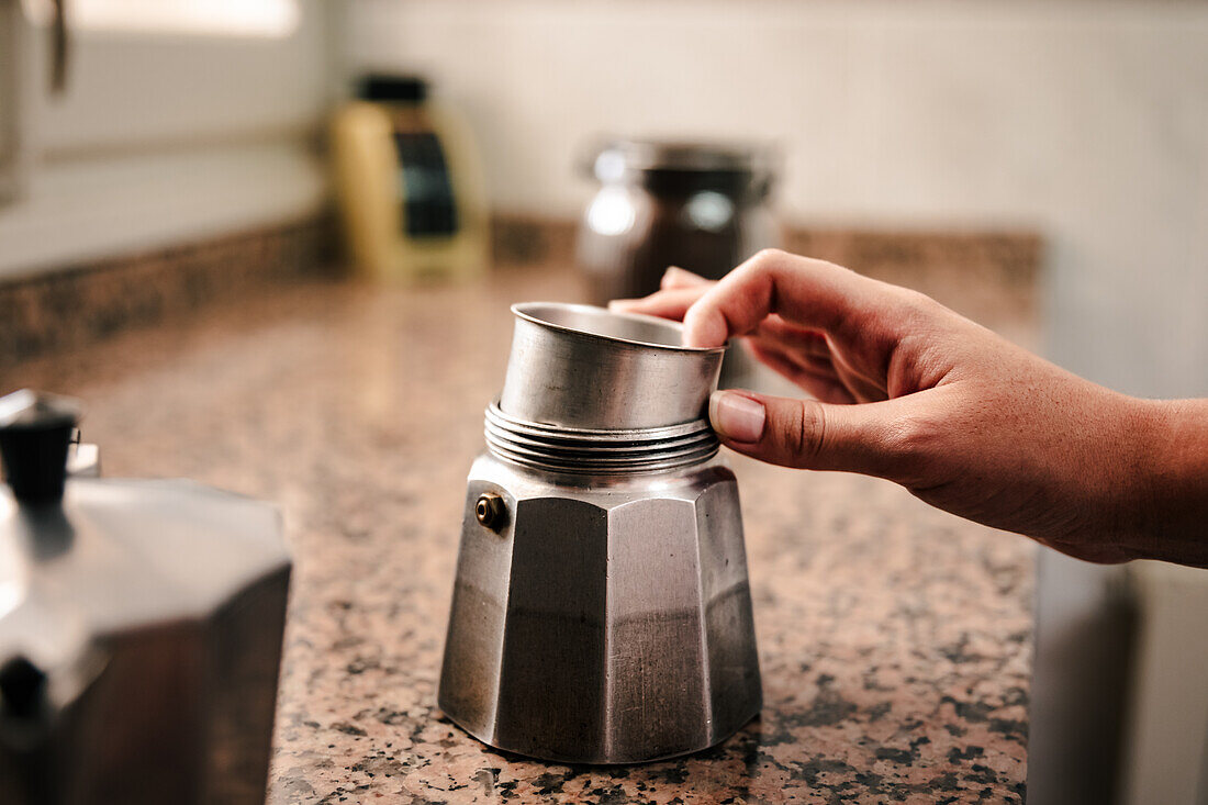 Nahaufnahme einer anonymen Hand, die eine italienische Mokkakanne auf einer Küchentheke zusammensetzt, mit unscharfem Küchengeschirr im Hintergrund, das auf die Zubereitung von Kaffee zu Hause hinweist