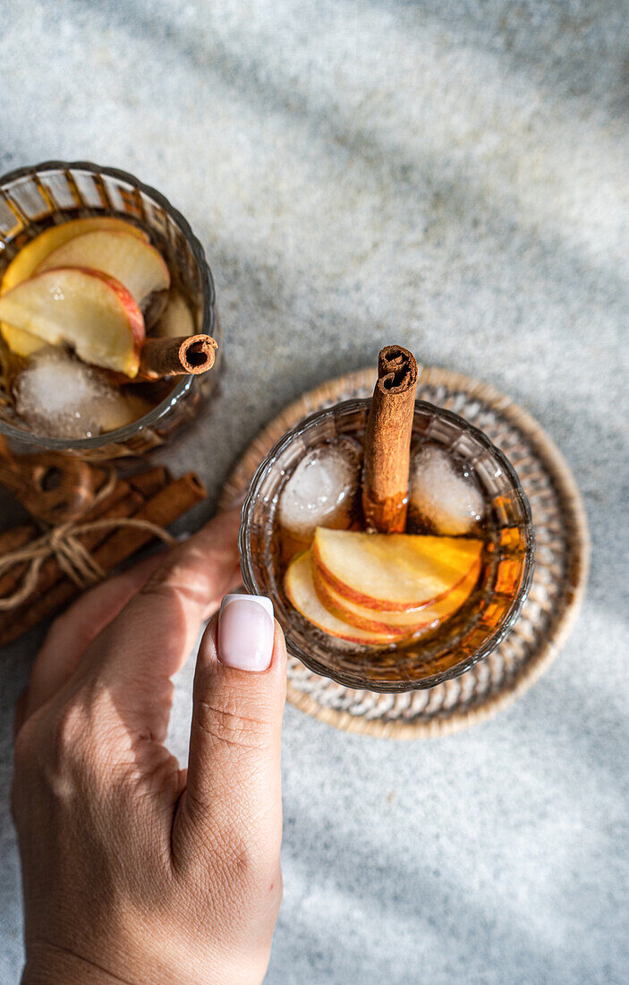 From above of anonymous hand holding a glass of apple cider cocktails adorned with cinnamon stick, star anise, and apple slices, showing fresh ice cubes