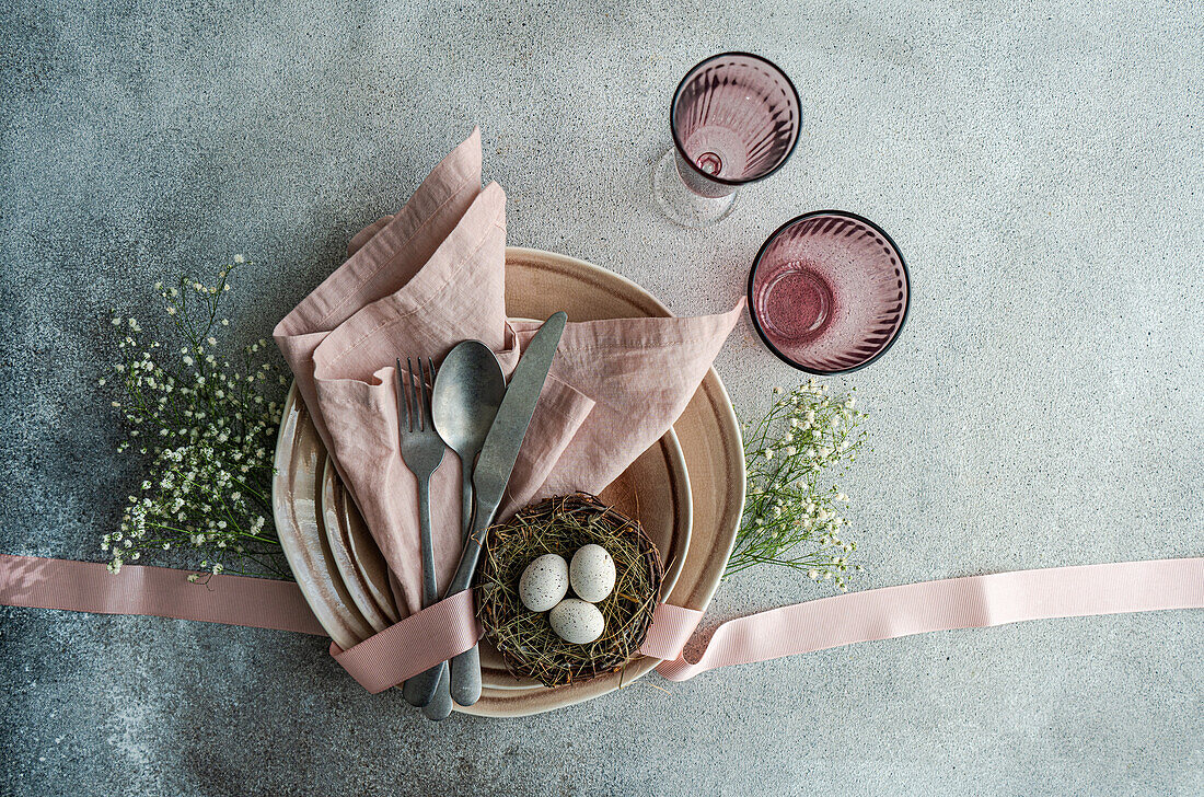 Draufsicht auf einen österlich gedeckten Tisch mit einem Nest mit Eiern, umgeben von Keramikgeschirr, Besteck und pastellfarbener Bettwäsche