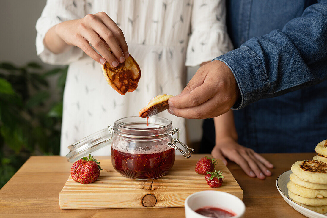 Crop anonymous persons in casual clothes standing at wooden table while eating delicious appetizing pancake with fresh strawberry jam on wooden board