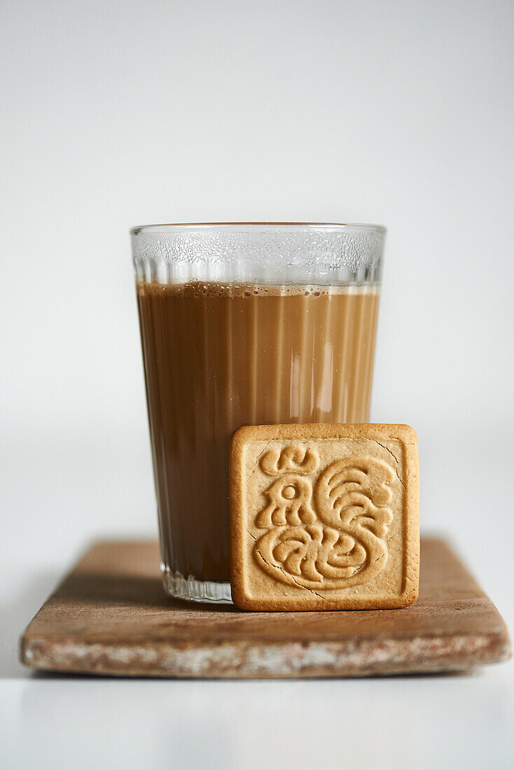 A cozy setting featuring a clear glass of warm tea latte, with a creamy froth on top, accompanied by a square biscuit with an intricate design, all resting on a rustic wooden coaster.