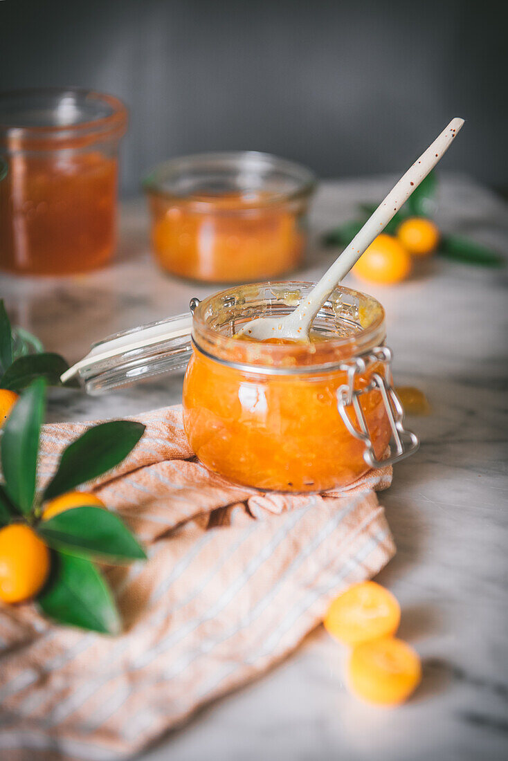 Nahaufnahme eines Glases mit frischer Kumquat-Marmelade mit Löffel auf einem Tisch vor verschwommenem dunkelgrauem Hintergrund