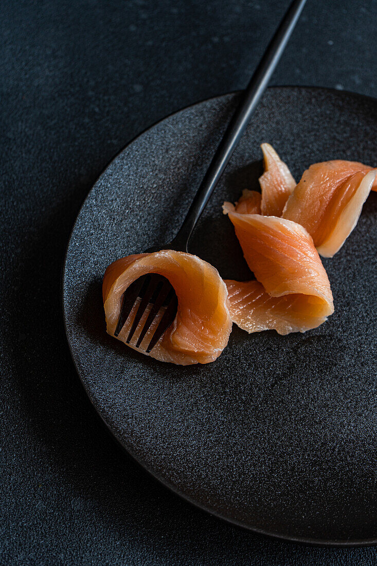 Top view of healthy salmon slice served on crop black plate near fork against dark surface