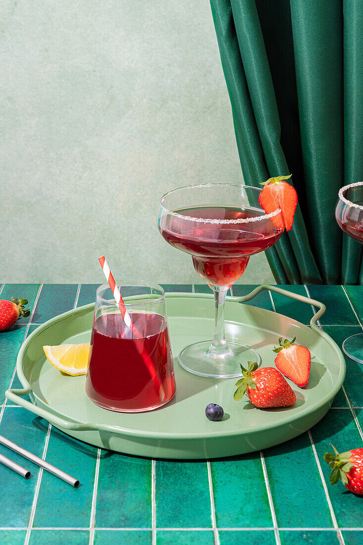 Gläser mit erfrischenden roten alkoholischen Cocktails mit Erdbeeren und Strohhalm, serviert auf einem Tablett mit Beeren und Zitronenscheibe auf dem Tisch