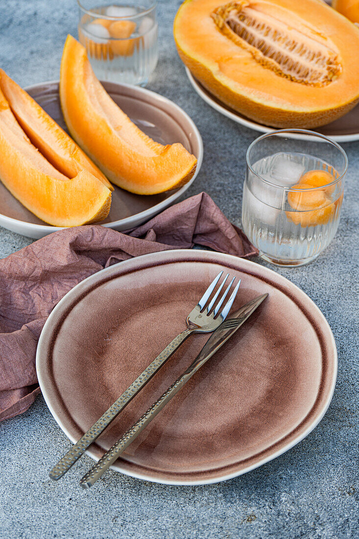 Hoher Winkel von in Scheiben geschnittener frischer Bio-Orangen-Moschus-Melone und Melonenkugeln in transparenten Gläsern mit Eiswürfeln, während sie auf dem Tisch mit Serviette, Gabel und Messer bei Tageslicht serviert werden