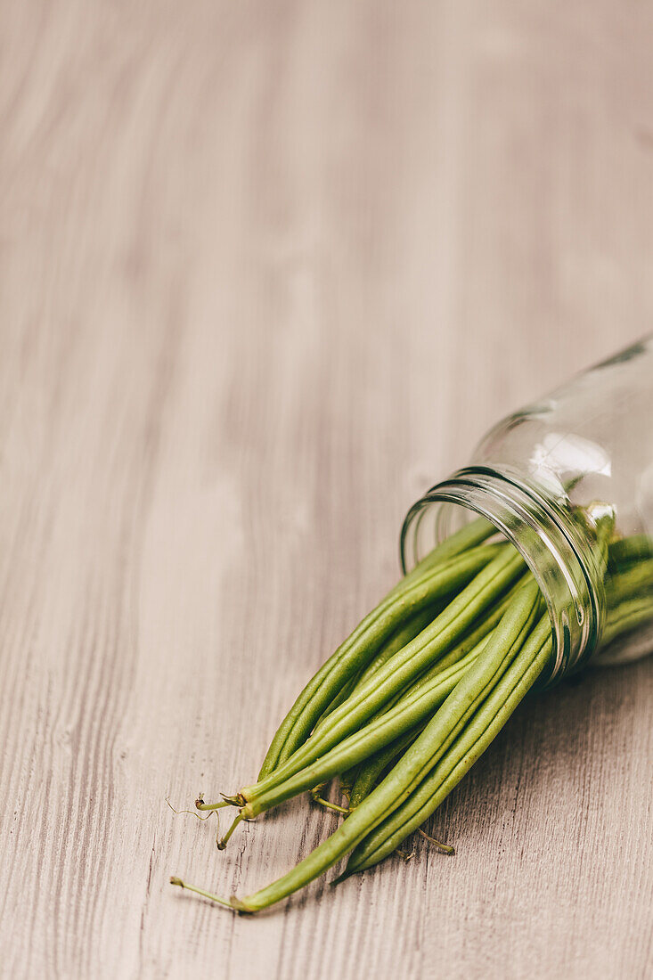 A clear glass jar tipped on its side, with fresh green beans cascading out onto a soft wooden surface.